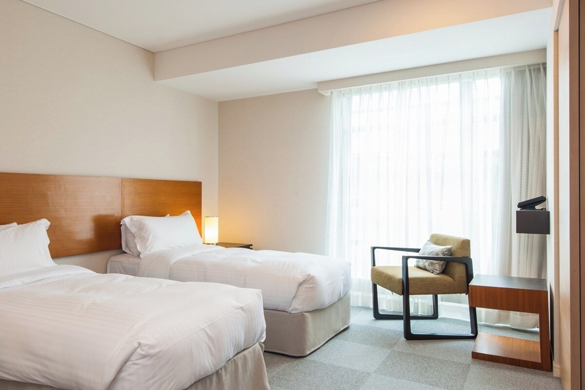 A hotel room with two beds, a desk, a chair, and a large window with curtains.