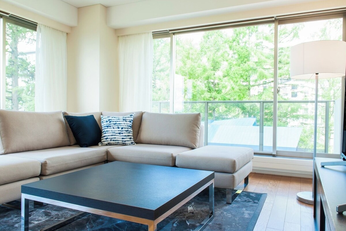 A modern living room with a beige sectional sofa, a dark coffee table, and large windows offering a view of trees and buildings.