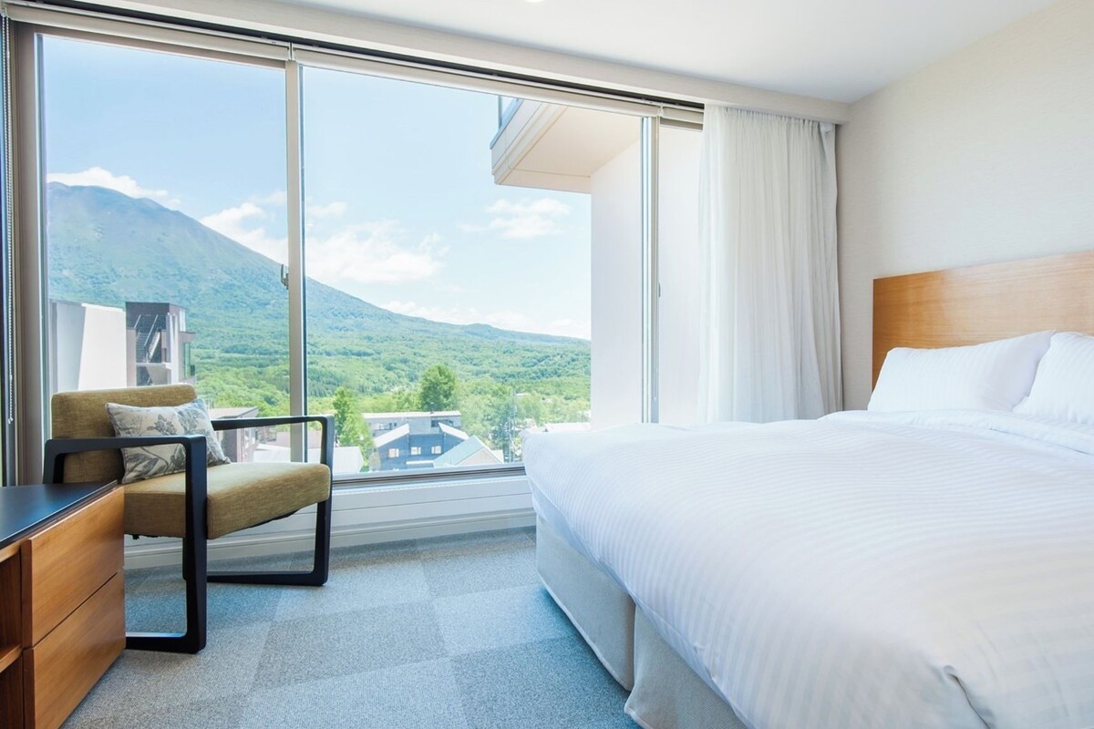 A hotel room with a large bed, a desk, and a chair. There is a window with a view of mountains and a city.