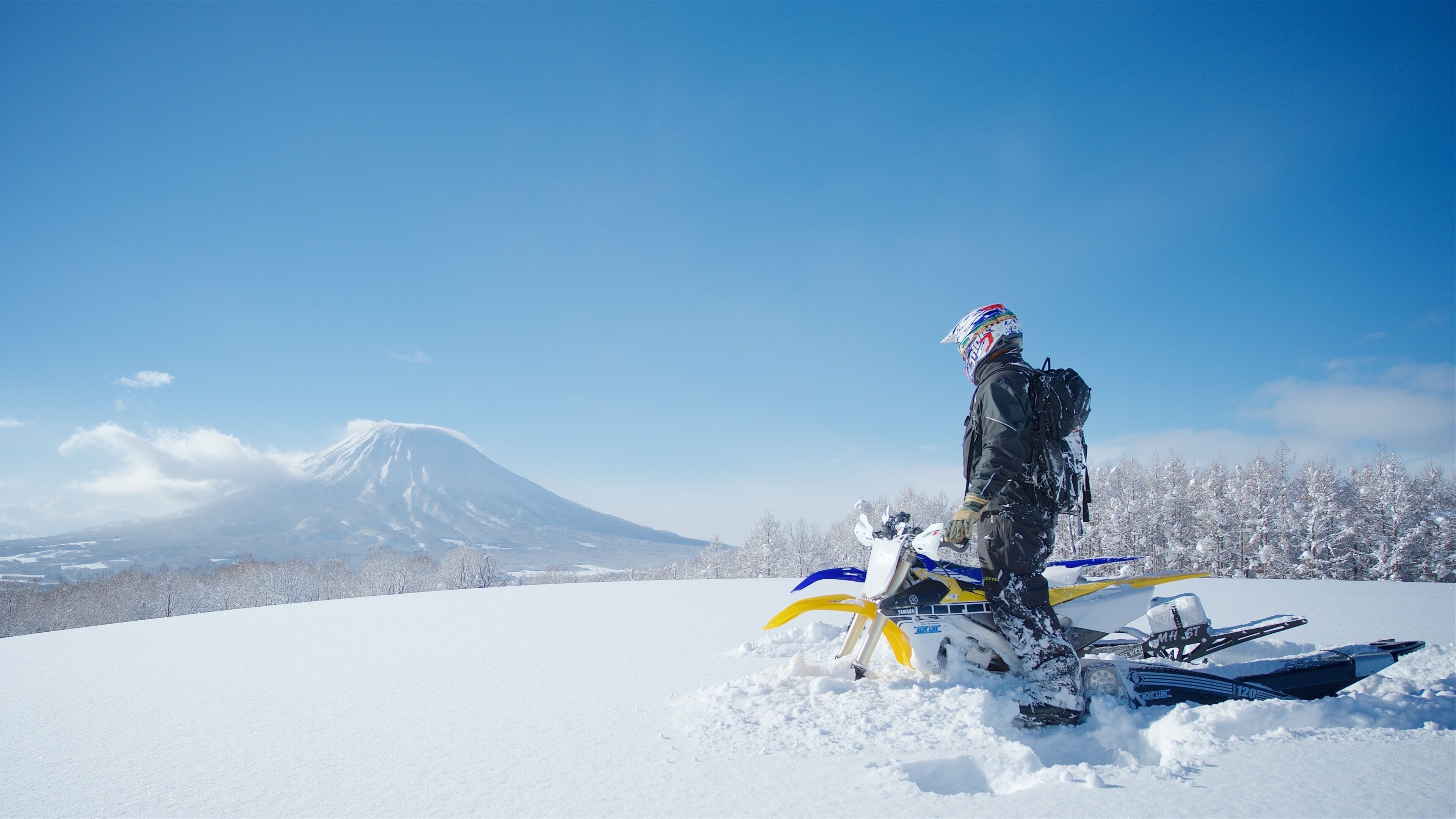 Un motoslitta con un conducente con abbigliamento invernale, un paesaggio innevato e una montagna sullo sfondo.