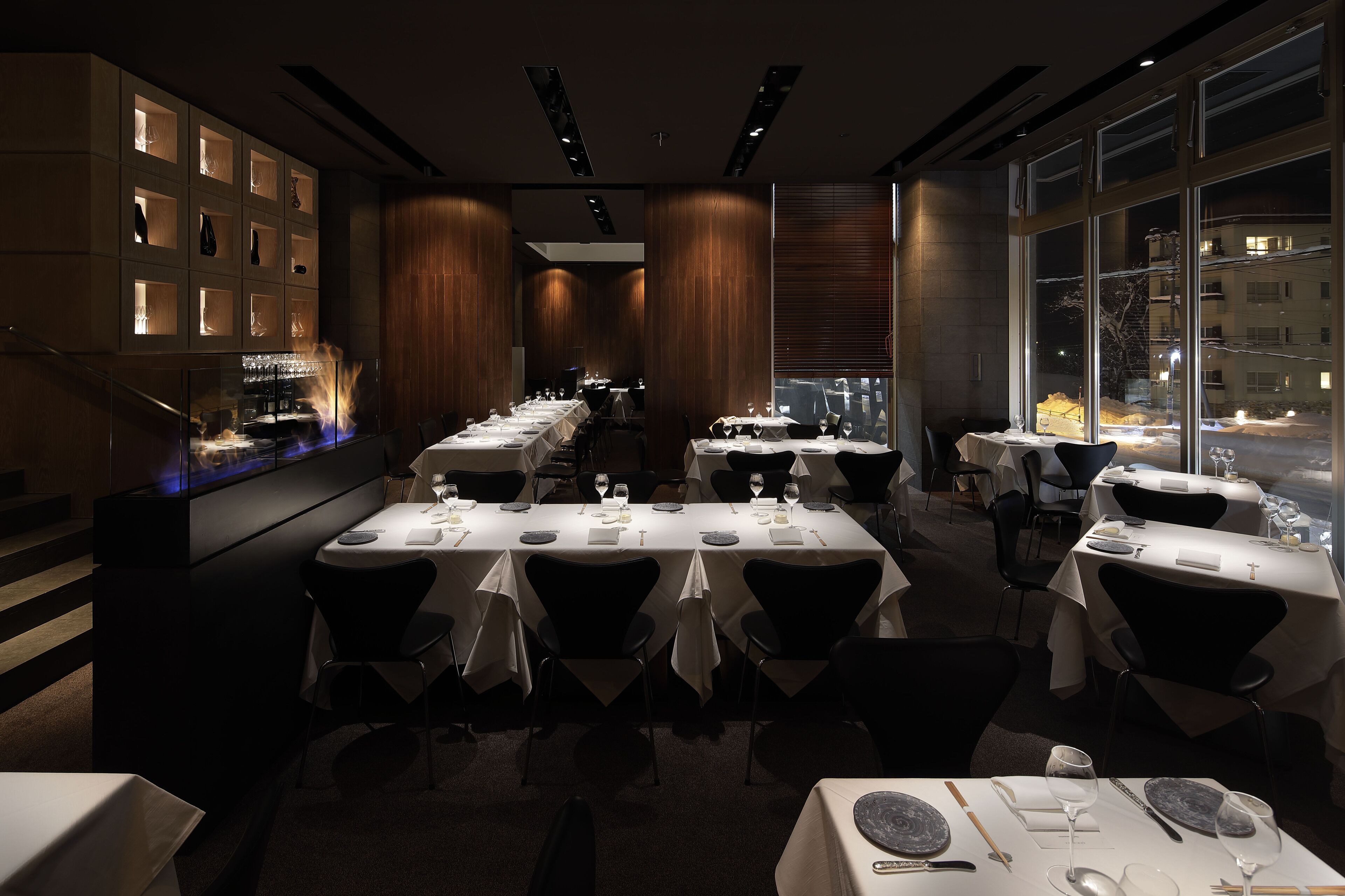 A restaurant with multiple tables set for dining, a bar area with illuminated shelves, and a view of the snowy cityscape through large windows.