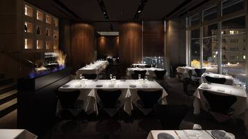 A restaurant with multiple tables set for dining, a bar area with illuminated shelves, and a view of the snowy cityscape through large windows.