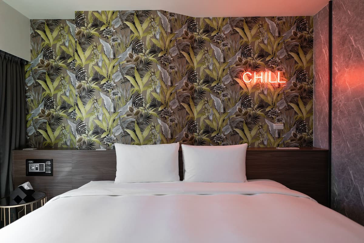 Habitación de hotel con cama, pared cubierta de papel tapiz floral, un letrero de neón que dice "CHILL" y un panel de control en la cabecera.