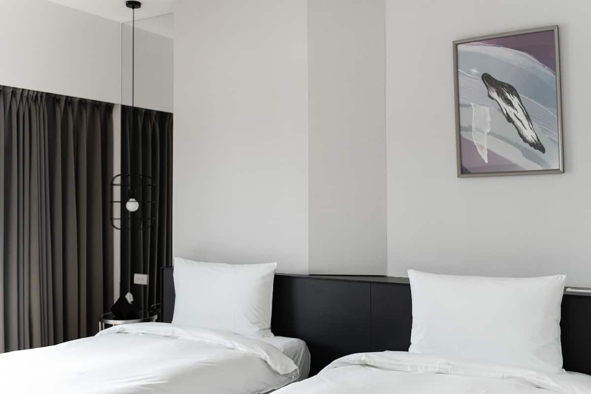 Una habitación de hotel moderna con dos camas, un ventanal con cortinas, una obra de arte colgada en la pared y una mesita de noche con lámpara.