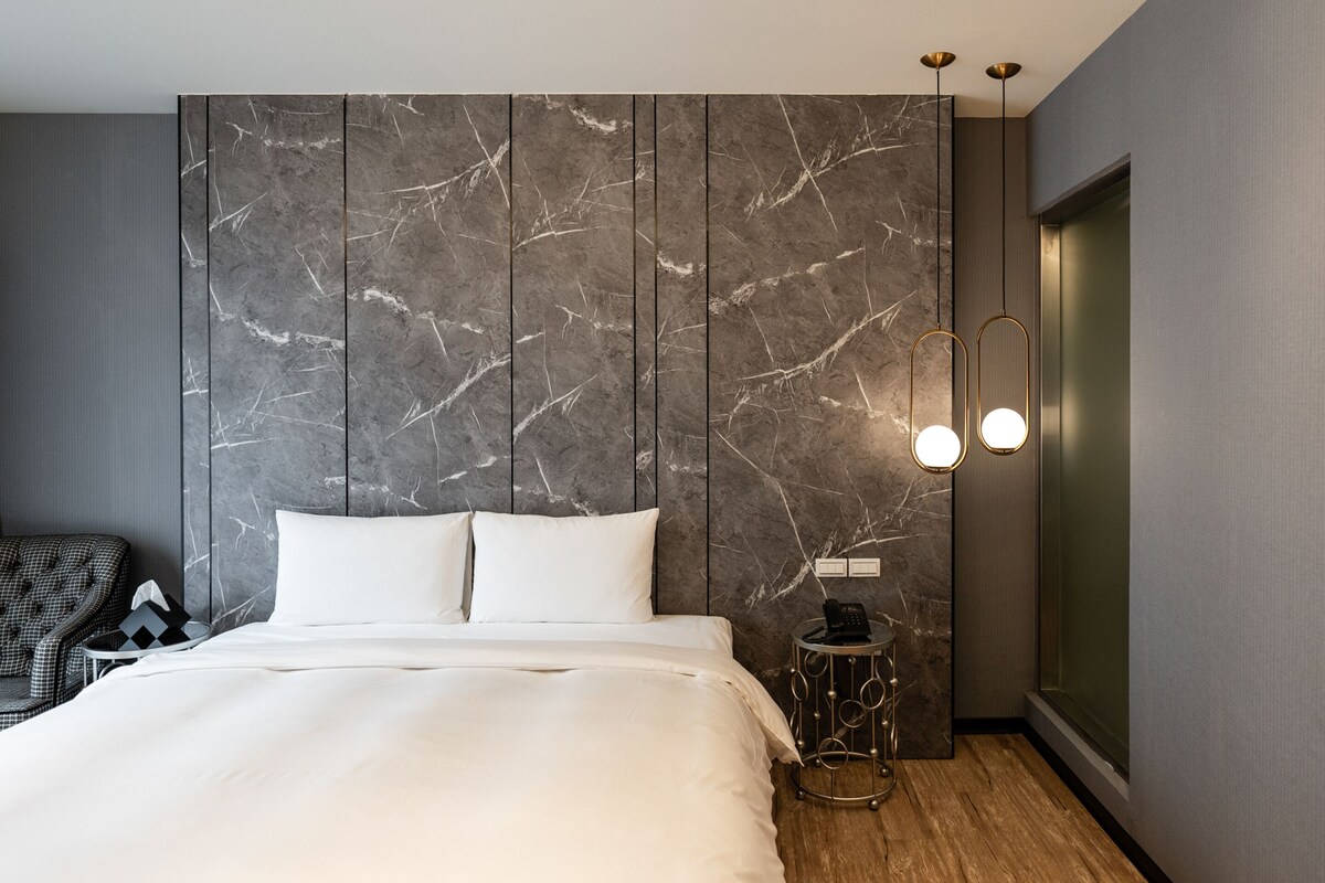 Een moderne slaapkamer met een groot bed, marmeren wandpanelen, een klein rond tafeltje met een telefoon en een stoel.
