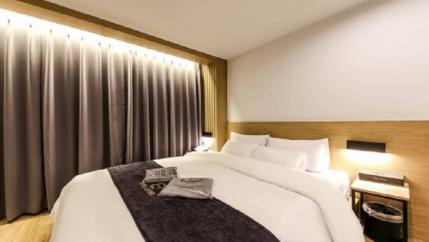 Habitació d'hotel amb un llit gran, un capçal, una tauleta de nit amb una làmpada i una finestra amb cortines.