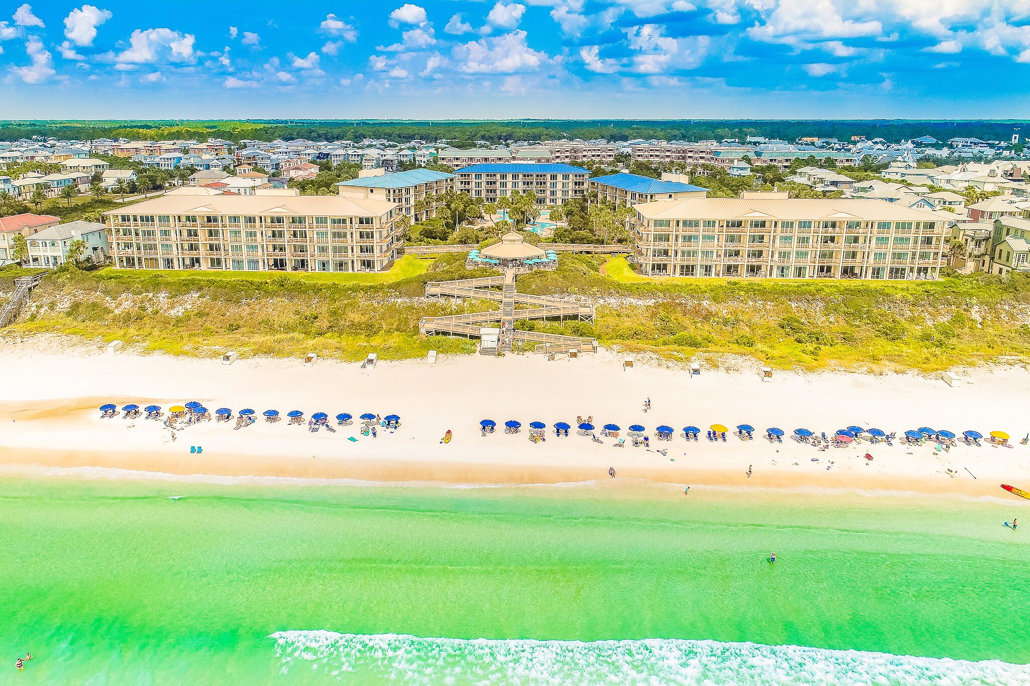 A coastal resort with multiple buildings, a beach with umbrellas, and a clear view of the ocean.