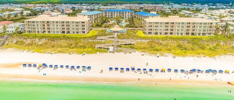 A coastal resort with multiple buildings, a beach with umbrellas, and a clear view of the ocean.