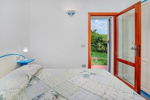 Quarto com cama, mesa de cabeceira, luminária na parede e porta para um jardim.