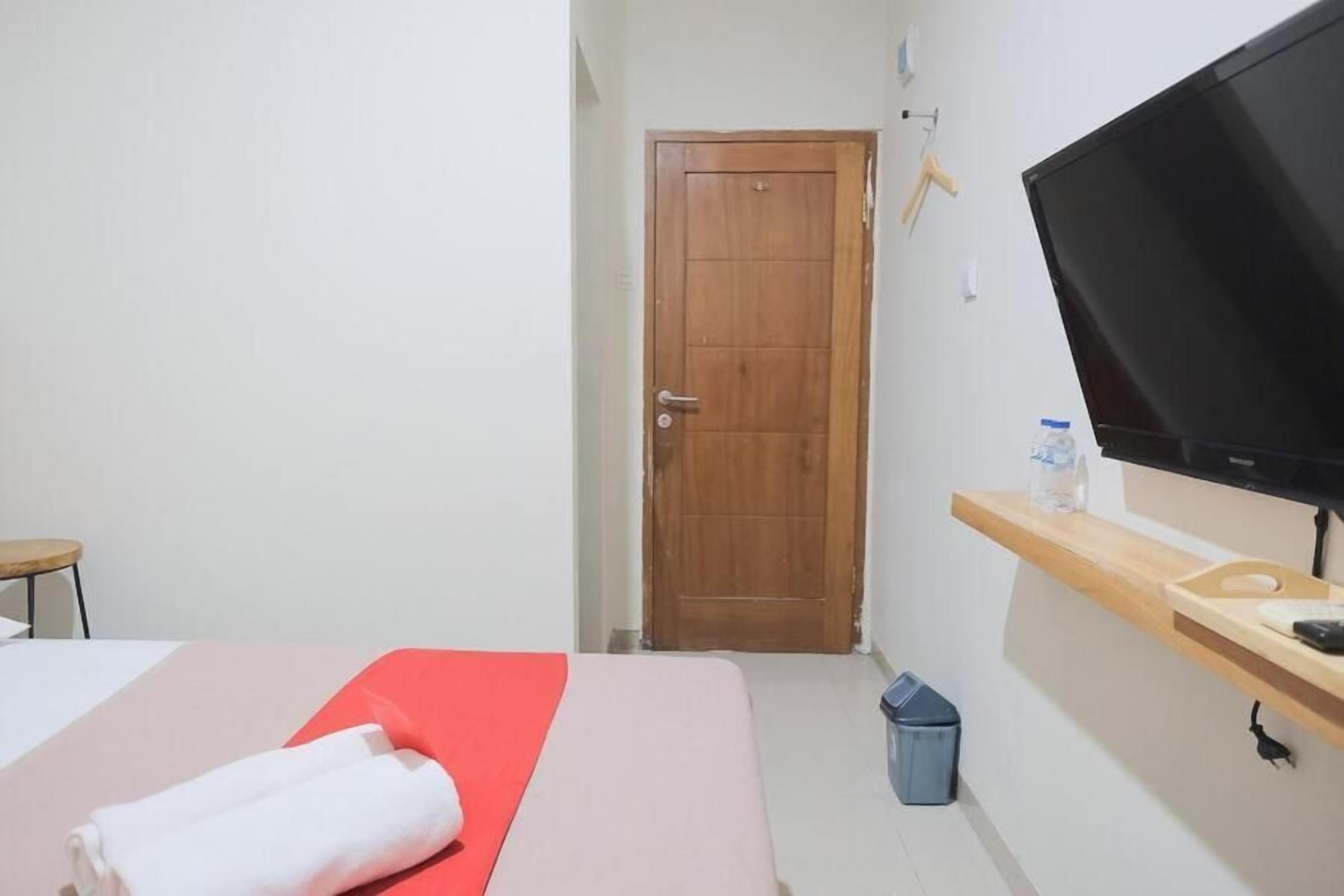 Habitación de hotel con cama, televisor, estante de madera con una botella y un cesto de basura.