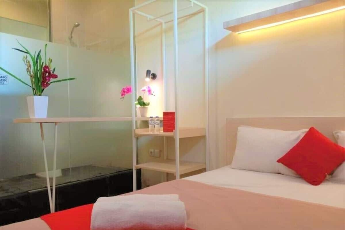 A hotel room with a bed, a red blanket, a white pillow, a red pillow, a small table with a plant, and a shelf with toiletries.