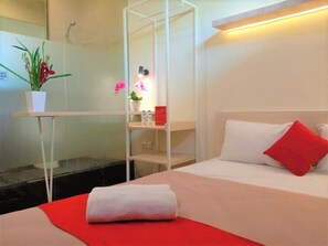 A hotel room with a bed, a red blanket, a white pillow, a red pillow, a small table with a plant, and a shelf with toiletries. - URVANA Hotel By Majestra Hospitality (Bogor)