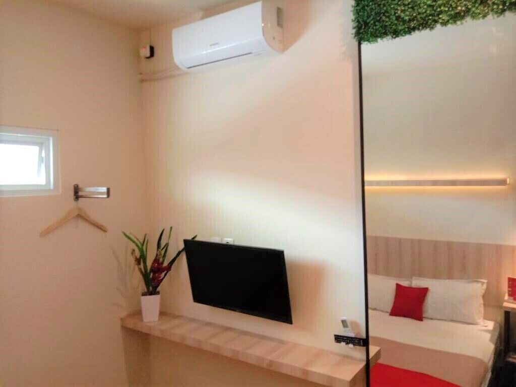 A hotel room with a mounted TV, a small plant, a bed with white and red pillows, and a window.