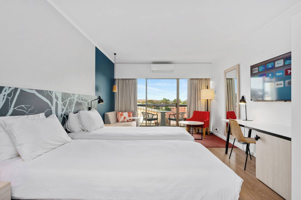 A hotel room with two beds, a desk, a chair, a TV, and a balcony with a table and chairs.