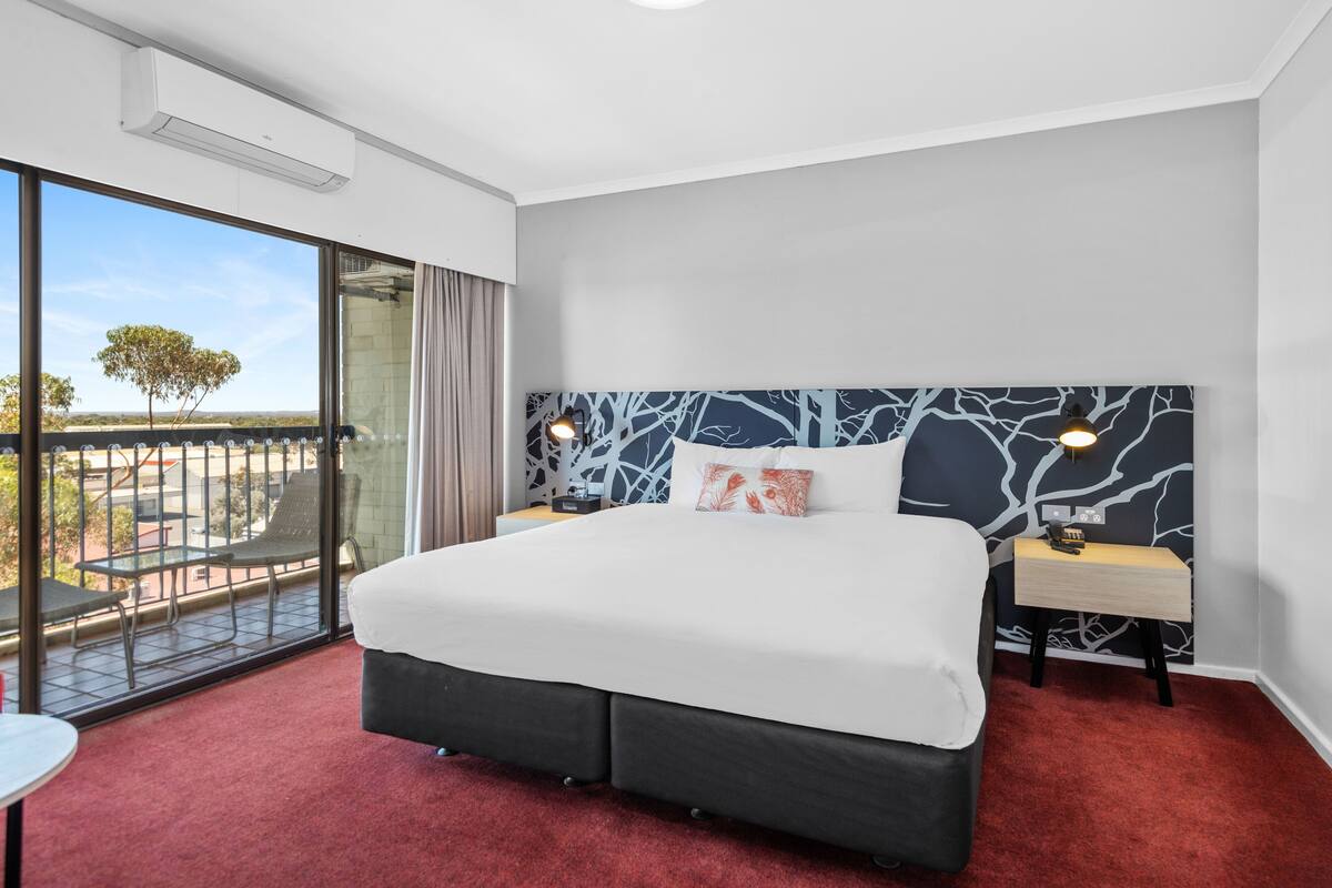 A hotel room with a large bed, a nightstand, a desk, a chair, and a balcony with a view.