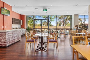 A spacious restaurant with wooden tables and chairs, a kitchen area, and large windows offering a view of the outdoors. - The Plaza Hotel Kalgoorlie (Kalgoorlie)