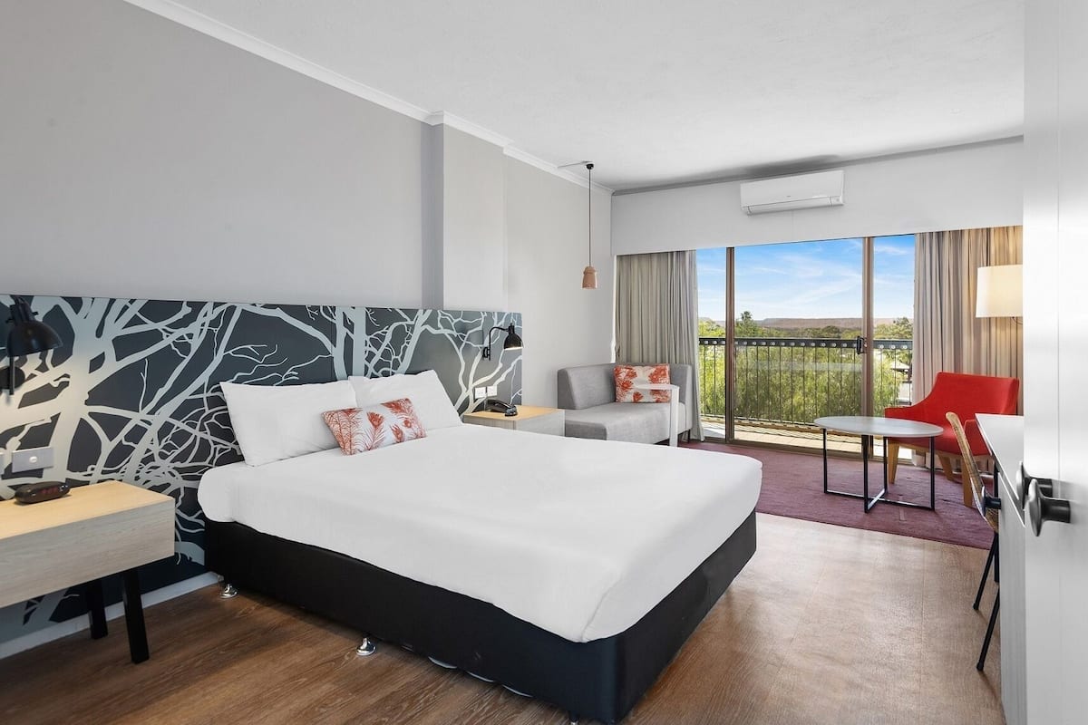 A hotel room with a large bed, a desk, a chair, and a balcony with a view.