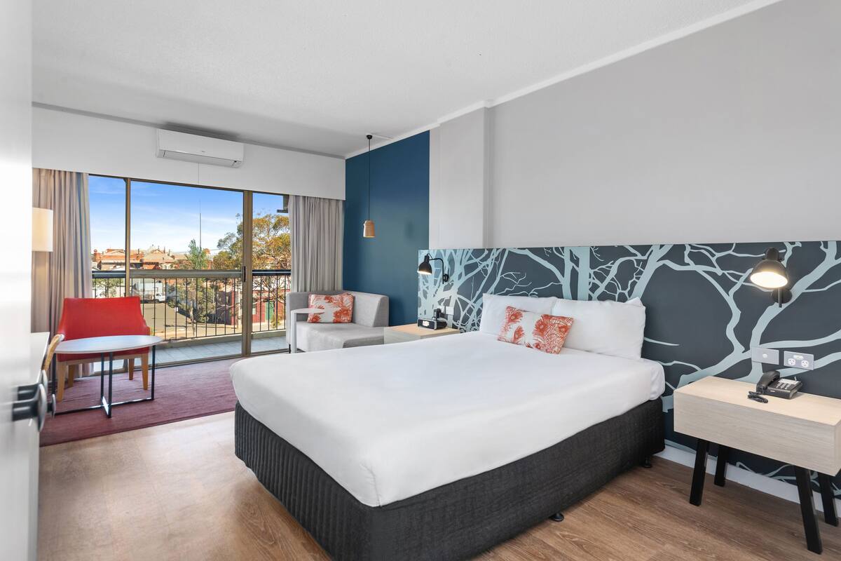 A modern hotel room with a large bed, a desk, a chair, and a view of the city.