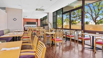 A spacious dining area with wooden tables and chairs, large windows offering a view of trees and buildings, and a modern ceiling with recessed lighting.