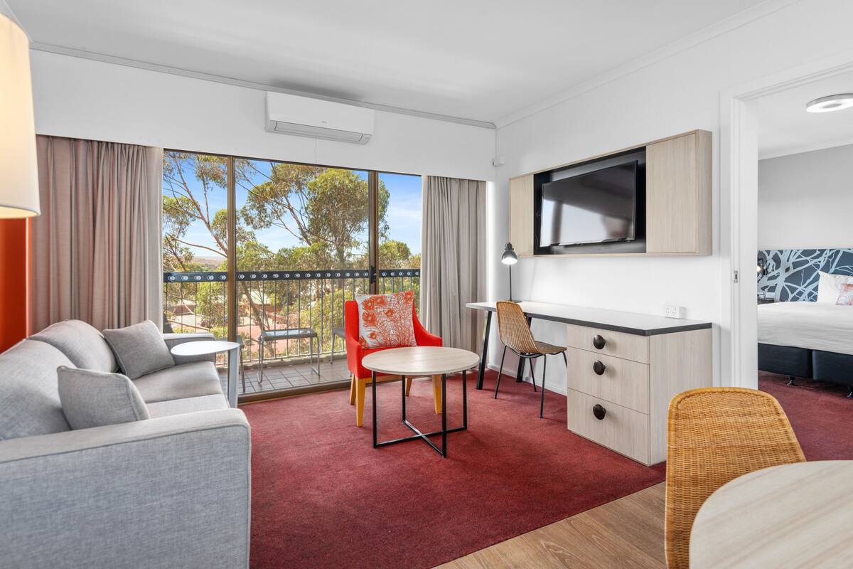 A modern hotel room with a sofa, dining table, chairs, a TV, and a bed.