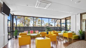Un espacio interior amplio con sillas y sofás amarillos dispuestos alrededor de una mesa central, ventanas grandes que ofrecen vistas a árboles y edificios, y un televisor de pantalla plana montado en la pared.