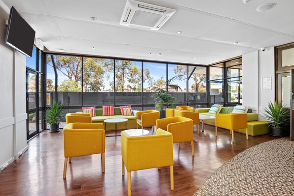 A spacious indoor area with yellow chairs and sofas arranged around a central table, large windows offering a view of trees and buildings, and a flat-screen TV mounted on the wall.