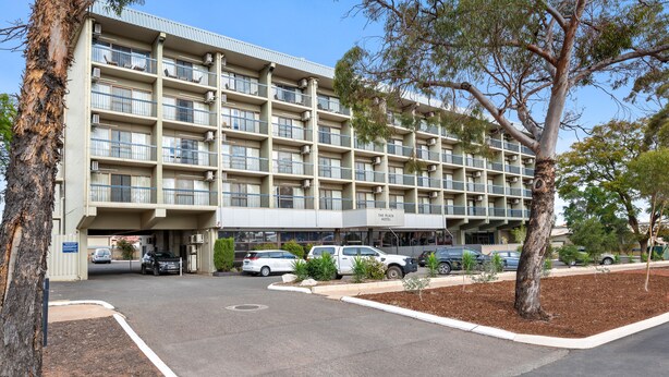 Un hotel de varios pisos con fachada plana, múltiples balcones y un estacionamiento con varios automóviles.