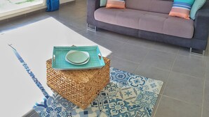 Ein Wohnzimmer mit grauem Sofa, bunten Kissen, einem Korbtisch mit Tellern und einem gemusterten Teppich. Durch eine Glastür ist der Blick in die Küche und den Außenbereich möglich.