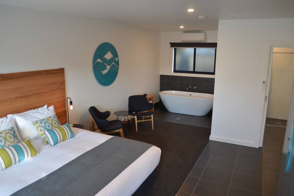 A modern hotel room with a large bed, a sitting area with a chair and a small table, a bathtub, and a large window.