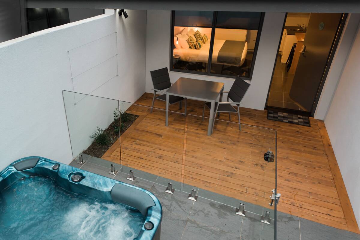 A modern outdoor patio with a hot tub, a dining table with chairs, and a view into a bedroom area.