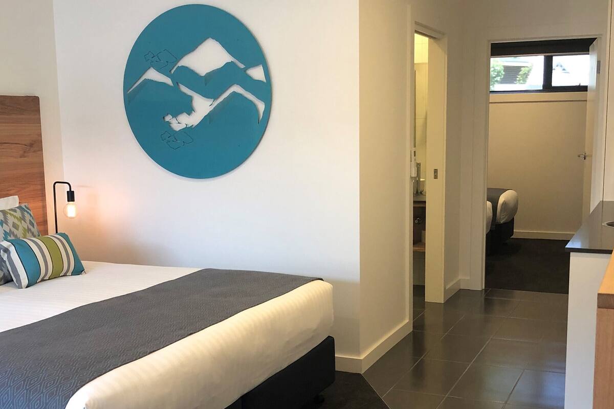 A modern hotel room with a bed, a wall-mounted light, a kitchen area, and a view through an open door.