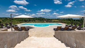 Zona de piscina con sillones de descanso y sombrillas, rodeada por un muro de piedra y vistas al campo.