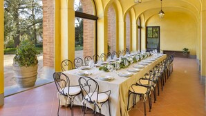 Una larga mesa de comedor dispuesta con sillas, rodeada de arcos y un muro de ladrillo, con una planta en maceta y una lámpara colgante.
