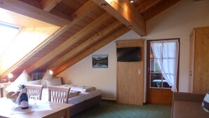 A cozy room with a sloped ceiling, a bed, a small table with a bottle and glass, and a TV mounted on the wall.
