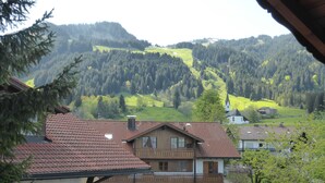 A traditional chalet with a sloped roof, a wooden balcony, and a view of a green valley and forested hills.