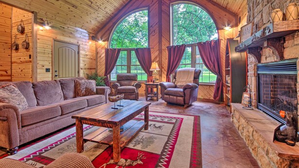 A cozy cabin interior with a wooden ceiling, a fireplace, a sofa, a coffee table, and two windows with curtains. - Spacious Family Home w/ Fire Pit by Norfork Lake! (Mountain Home)