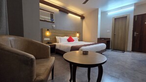 A hotel room with a bed, a chair, a small table, a bedside table with a lamp, a wall-mounted air conditioner, and a door. - Hotel Gulmohar Grand (East Jorhat)
