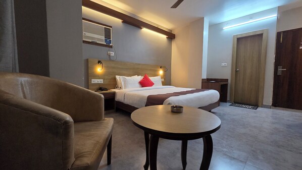 A hotel room with a bed, a chair, a small table, a bedside table with a lamp, a wall-mounted air conditioner, and a door. - Hotel Gulmohar Grand (East Jorhat)