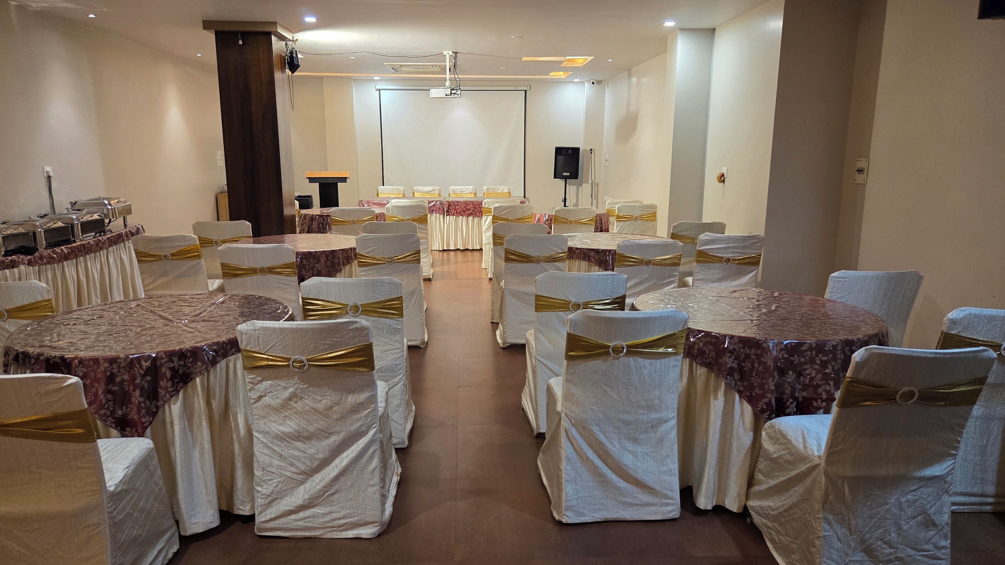 A banquet hall with round tables, chairs with white covers and gold sashes, a stage with a podium, and a projector screen.