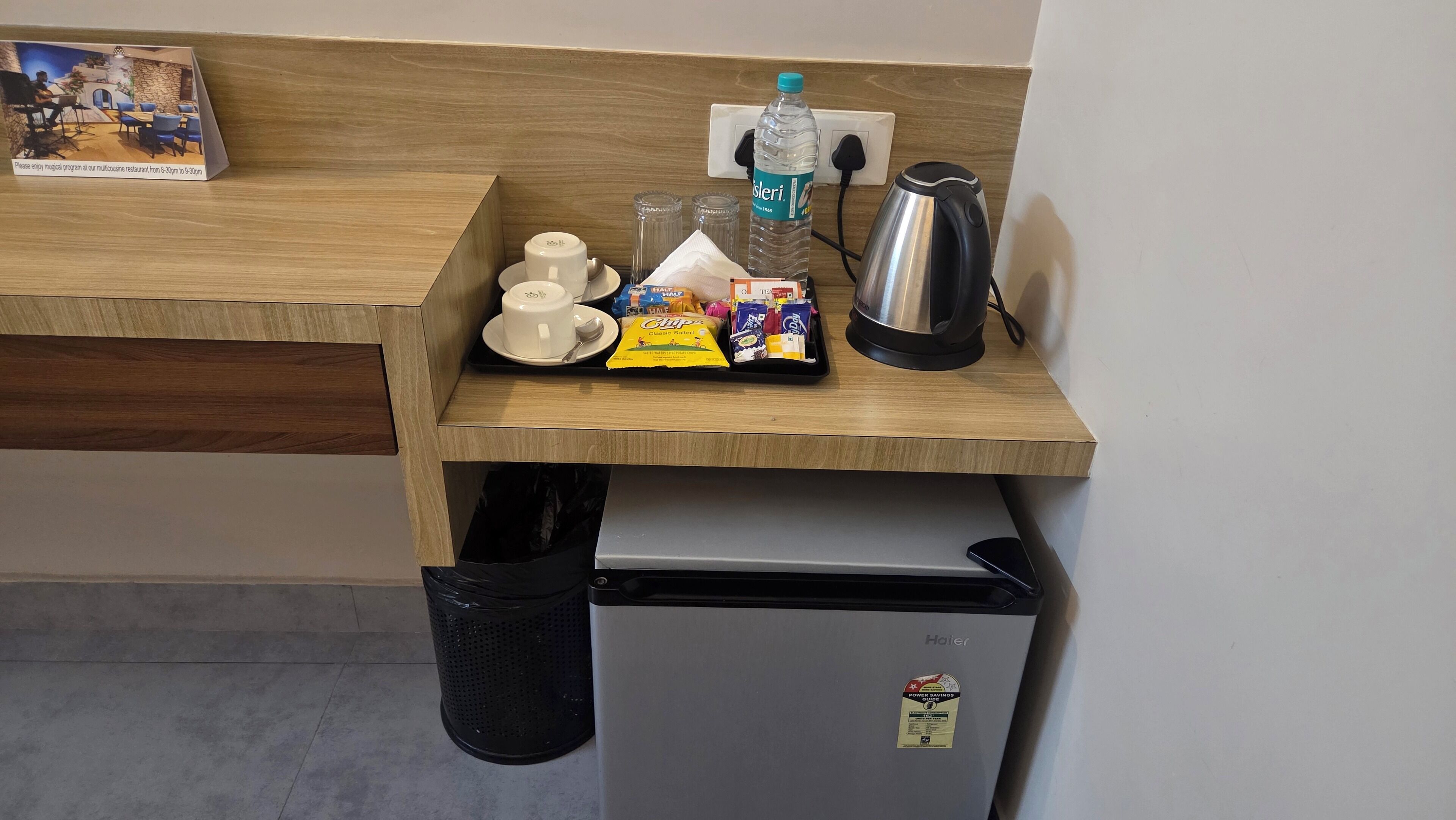 A kitchenette with a coffee maker, a kettle, a bottle of water, and a trash bin.