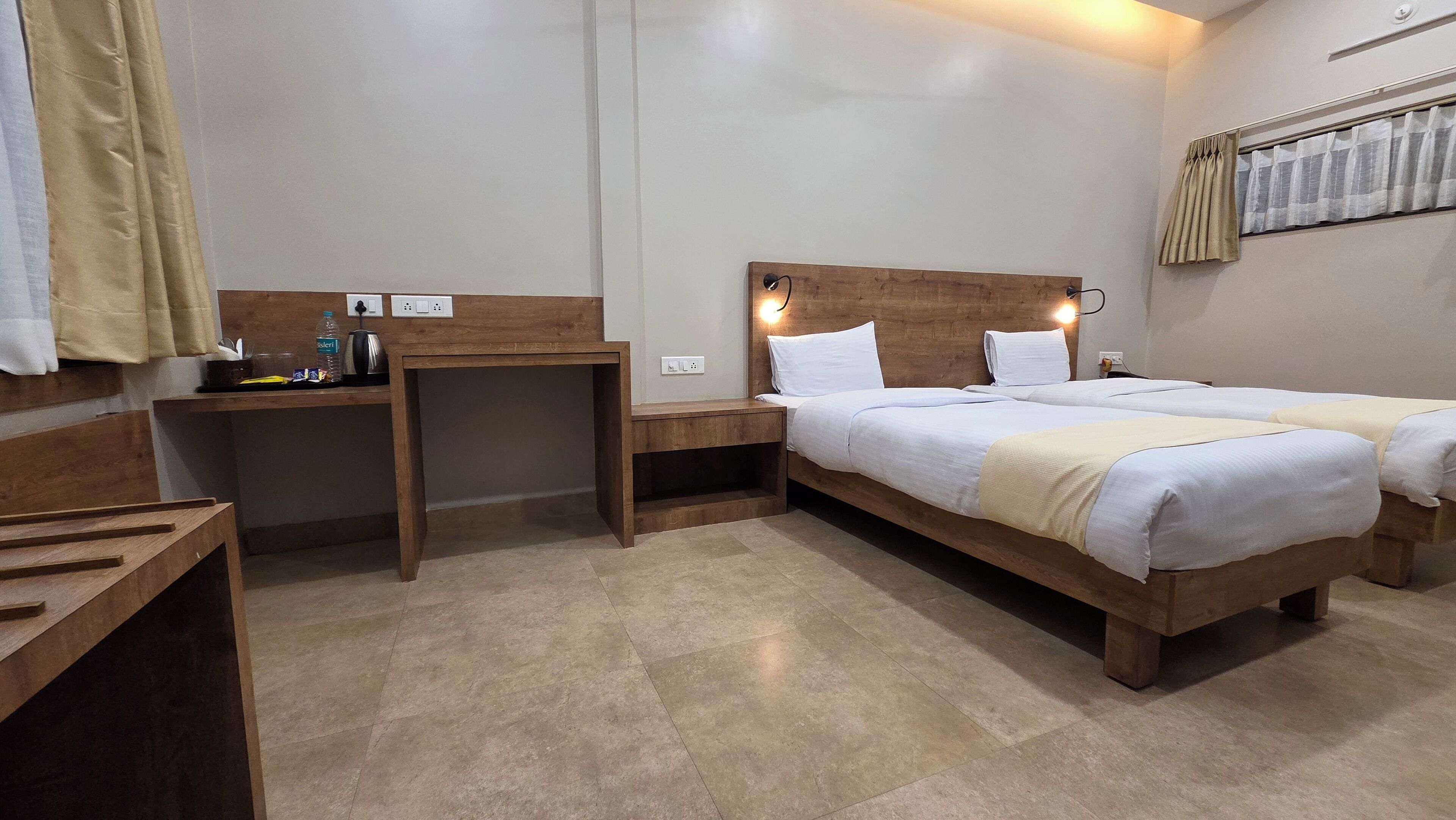 A hotel room with a wooden bed, bedside tables, a desk, and a small table with a bottle and a kettle.
