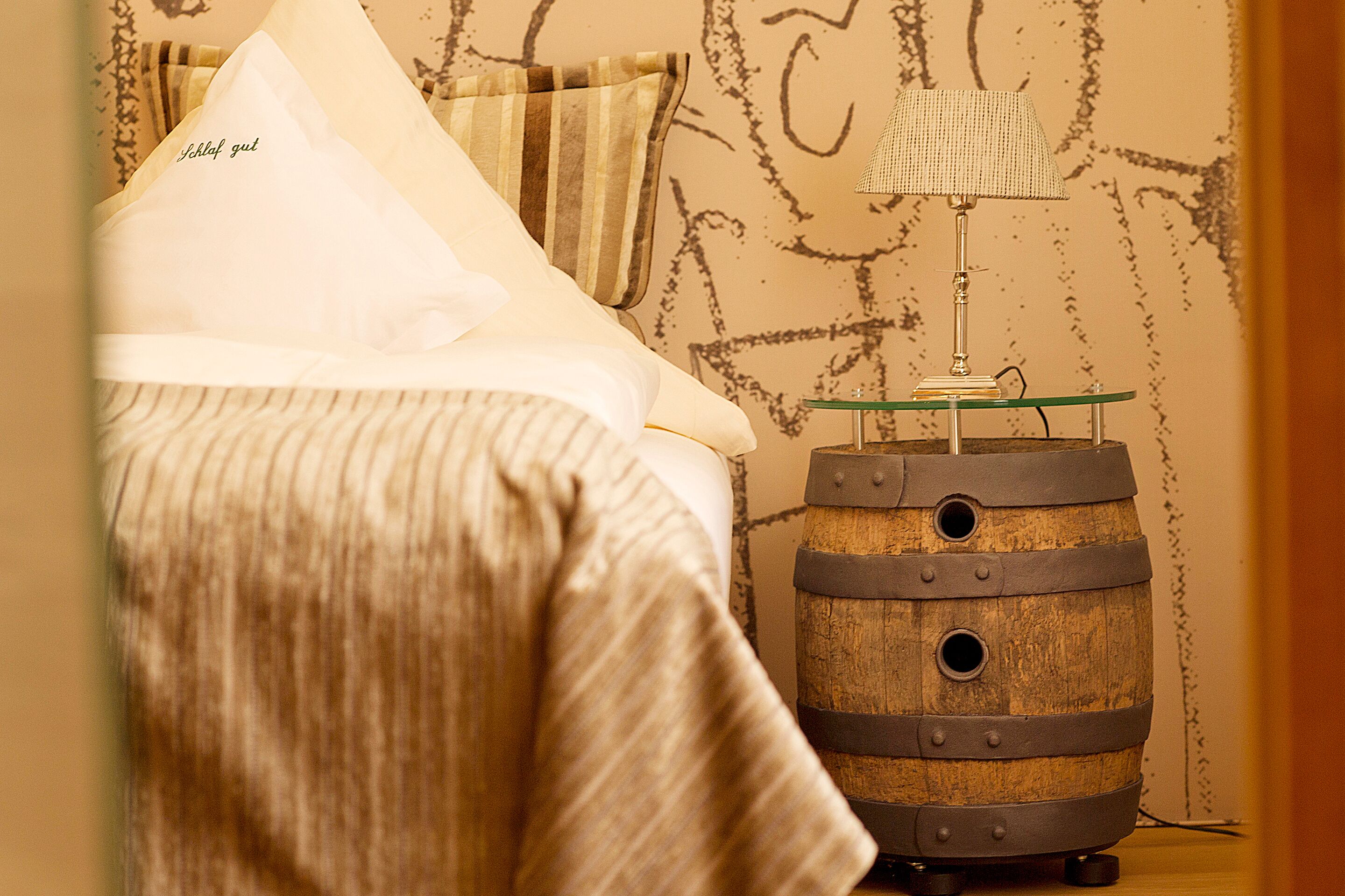A bed with a pillow labeled "Schlaf gut," a striped blanket, and a wooden barrel with a lamp on top.