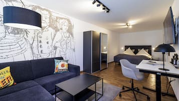 A modern living room with a sofa, coffee table, and a desk with a computer. There is a bed with a headboard and a TV mounted on the wall.