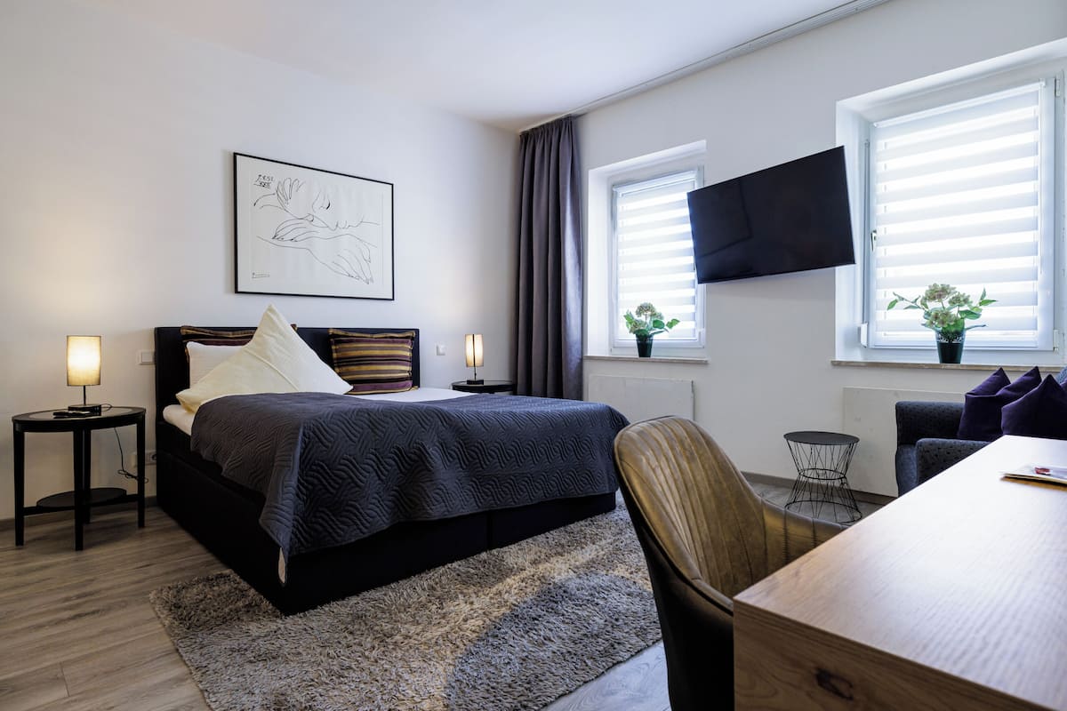 A modern hotel room with a bed, a desk, a chair, a TV, and a window with blinds.