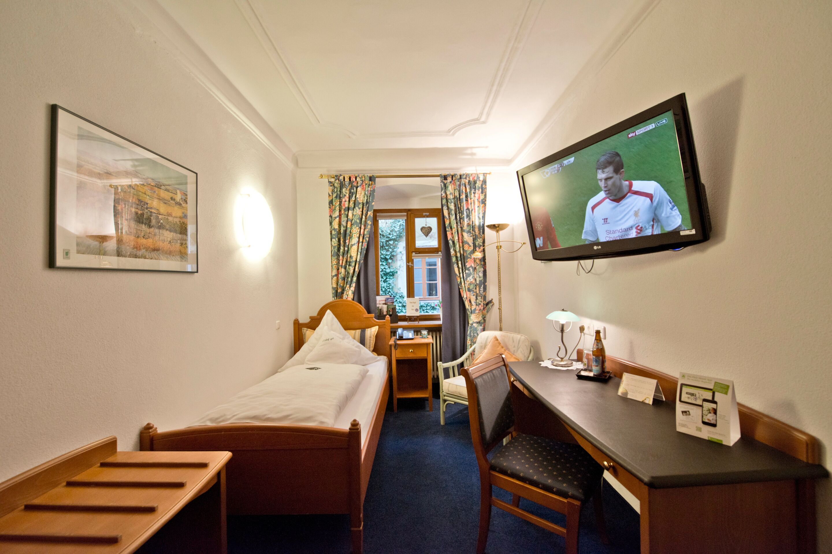 Quarto de hotel com cama, escrivaninha, cadeira, televisão e janela com cortinas.