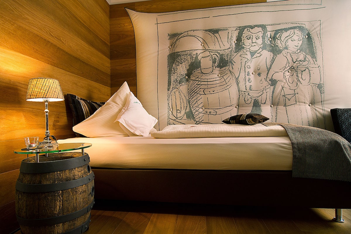 A bed with a large tapestry, a wooden barrel lamp, and a glass table.