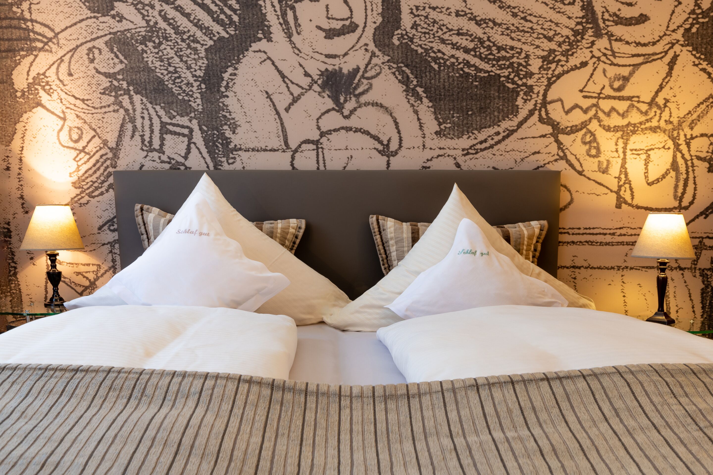 Una cama bien tendida con ropa de cama blanca y cubrecamas a rayas, flanqueada por dos lámparas de noche y un cabecero con diseño estampado.