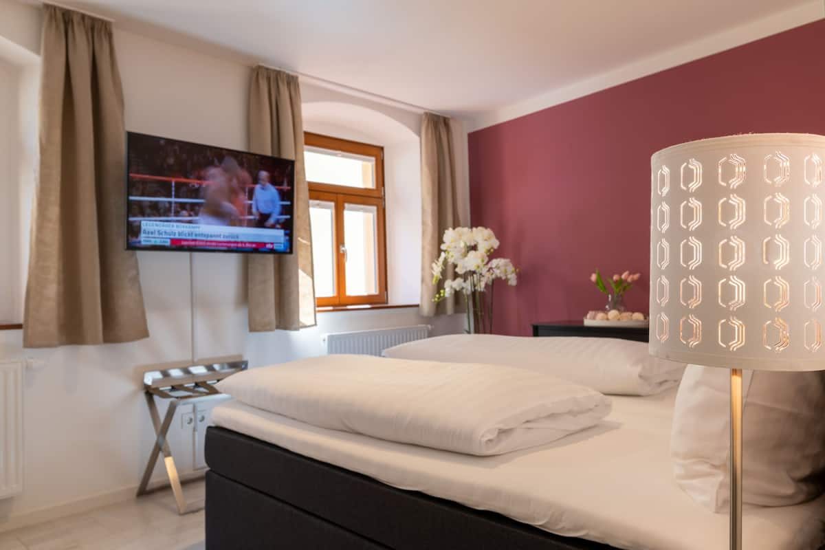 A hotel room with a bed, a TV mounted on the wall, a window with curtains, and a bedside table with a lamp.