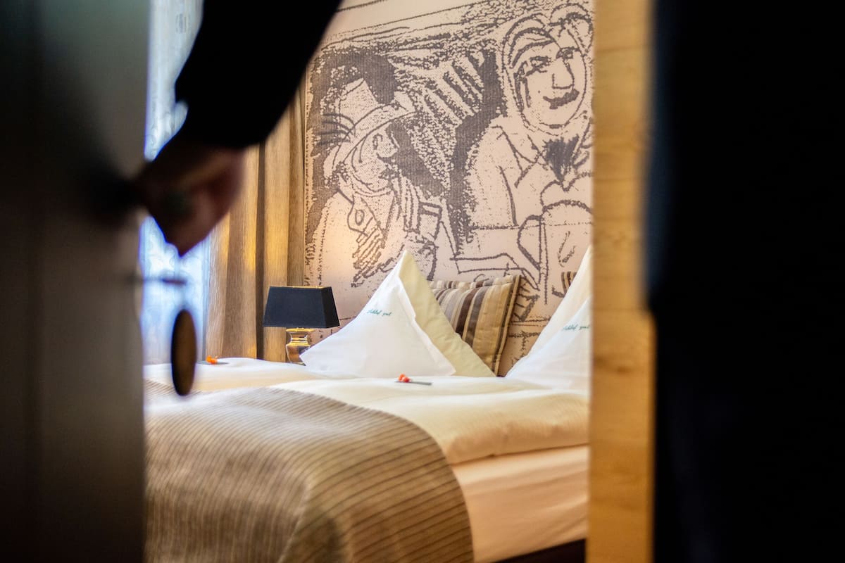 A hotel room with a bed, pillows, a nightstand, and a wall tapestry.