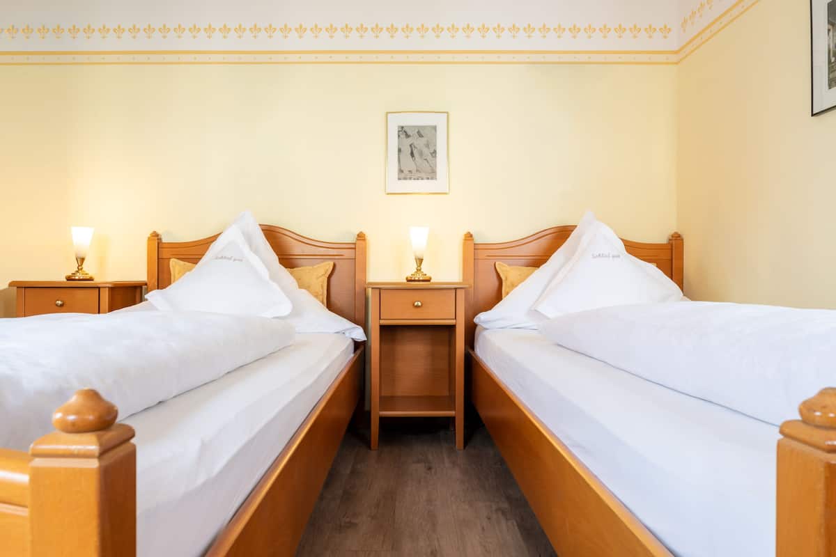 A hotel room with two wooden beds, white linens, nightstands with lamps, a framed picture on the wall, and a wooden floor.